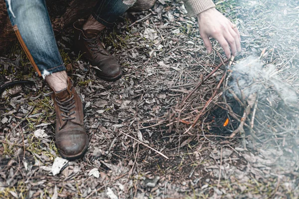 A stock photo of a man in nature sitting by a small fire, warming himself.