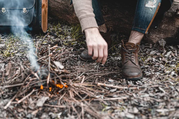 A stock photo of a man in nature sitting by a small fire, warming himself.