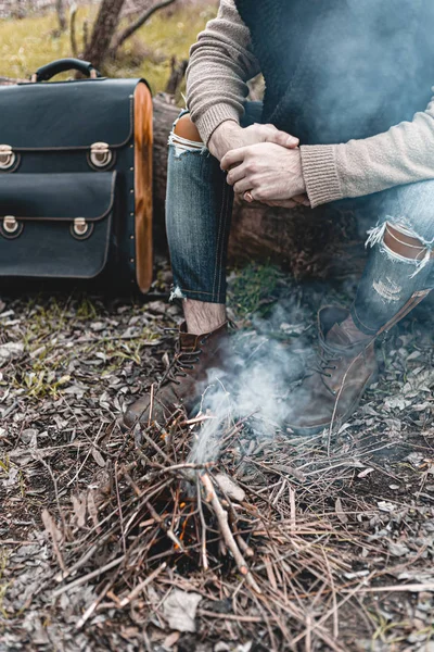 A stock photo of a man in nature sitting by a small fire, warming himself.