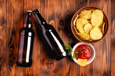 Two glass bottles of beer without a label with a snack in the form of potato chips with red sauce. Stock photo beer with snack on a wooden table, top view.