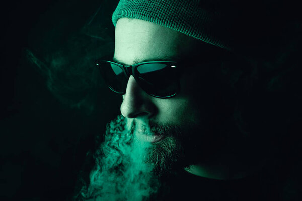 Portrait of a bearded, white hipster smoker on a dark background. Studio stock photo concept of smoking cigarettes and marijuana.