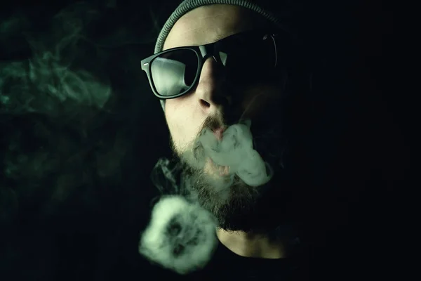 Portrait of a bearded, white hipster smoker on a dark background. Studio stock photo concept of smoking cigarettes and marijuana.