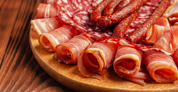 Assorted meat snacks on a wooden cutting board. Sausage, ham, bacon, smoked meats. Stock photo of meat products with blank space.