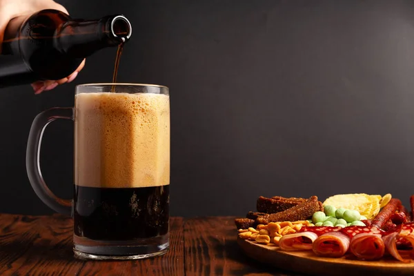 木製のテーブルの上にビールと軽食の品揃えのマグカップ ソーセージ サラミ ベーコン ビールの塩味のスナック チップ わさびナッツ 塩ピーナッツ クルトン — ストック写真