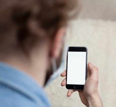 Adam evde tıbbi bir maskenin içinde boş bir akıllı telefon görüntüsü gösteriyor. Coronavirus karantinası kavramını ve telefon başvurusunu kabul et veya tıbbi servisi ara.