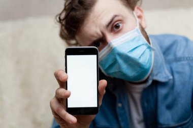 Adam evde tıbbi bir maskenin içinde boş bir akıllı telefon görüntüsü gösteriyor. Coronavirus karantinası kavramını ve telefon başvurusunu kabul et veya tıbbi servisi ara.