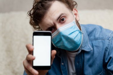 Adam evde tıbbi bir maskenin içinde boş bir akıllı telefon görüntüsü gösteriyor. Coronavirus karantinası kavramını ve telefon başvurusunu kabul et veya tıbbi servisi ara.