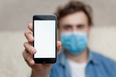 Adam evde tıbbi bir maskenin içinde boş bir akıllı telefon görüntüsü gösteriyor. Coronavirus karantinası kavramını ve telefon başvurusunu kabul et veya tıbbi servisi ara.
