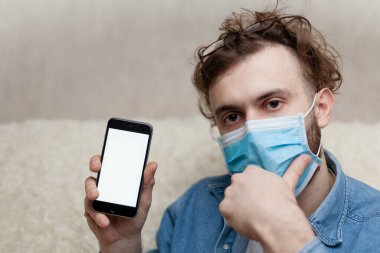 Adam evde tıbbi bir maskenin içinde boş bir akıllı telefon görüntüsü gösteriyor. Coronavirus karantinası kavramını ve telefon başvurusunu kabul et veya tıbbi servisi ara.