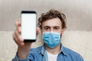 Adam evde tıbbi bir maskenin içinde boş bir akıllı telefon görüntüsü gösteriyor. Coronavirus karantinası kavramını ve telefon başvurusunu kabul et veya tıbbi servisi ara.