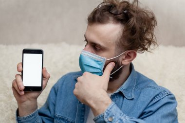 Adam evde tıbbi bir maskenin içinde boş bir akıllı telefon görüntüsü gösteriyor. Coronavirus karantinası kavramını ve telefon başvurusunu kabul et veya tıbbi servisi ara.