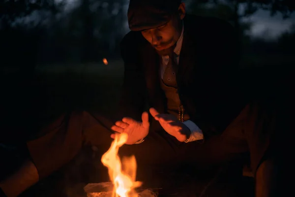 一个人在自然界的夜晚坐在火边的画像 电影老式照片 20世纪20年代的肖像画 一个戴着平顶帽子的英国人 — 图库照片