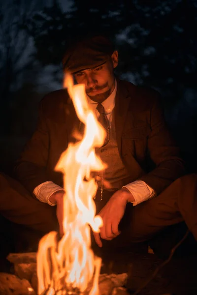 一个人在自然界的夜晚坐在火边的画像 电影老式照片 20世纪20年代的肖像画 一个戴着平顶帽子的英国人 — 图库照片
