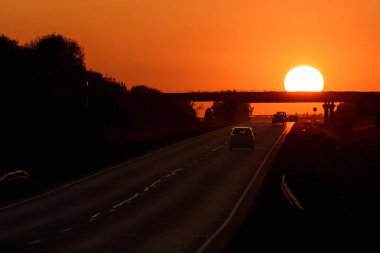 Bir asfalt yol, arabalar ve yuvarlak bir güneş üzerinde bir köprü bir ateşli kırmızı-turuncu gökyüzü günbatımı, arka planı renkli arka plan