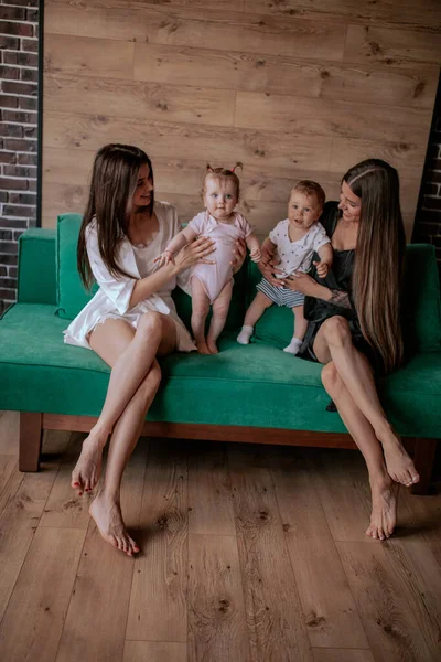 Dos atractivas madres en hermosos pijamas juegan con sus hijos en el sofá por la mañana —  Fotos de Stock