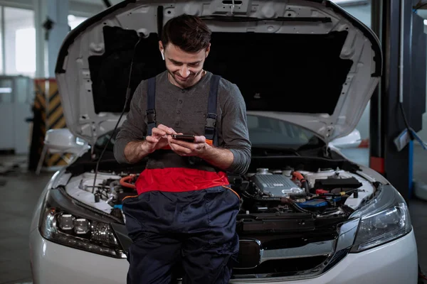 In the garage mechanic guy very attractive listening music from his wireless headphones and take a look on his phone while have the break time