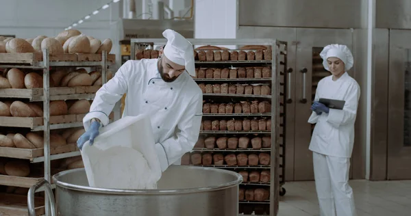 Bakery industry big chef in a special uniform preparing the dough add some flour from the basket bakery engineer monitoring all the process background baked bread on the shelves