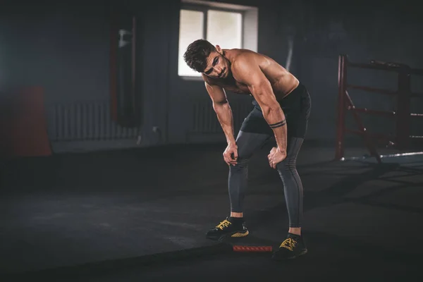 Spor giyim bölümündeki karizmatik adam spor salonunda dizlerinin üzerine çökmüş. Spor salonundaki adam yoğun idmandan sonra yorgun görünüyor.