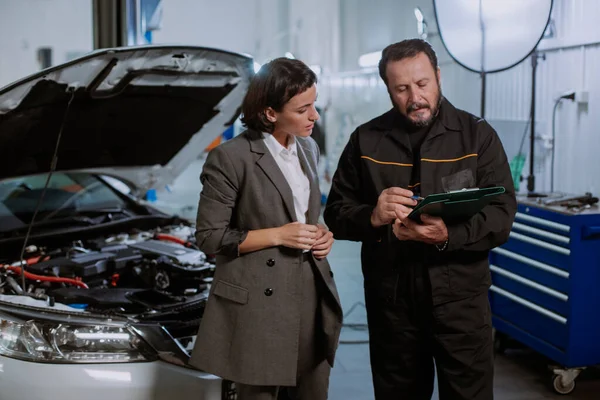 In a big service auto center charismatic mechanic man in a uniform with a businesswoman sign some papers then they come beside of the car to check the problem