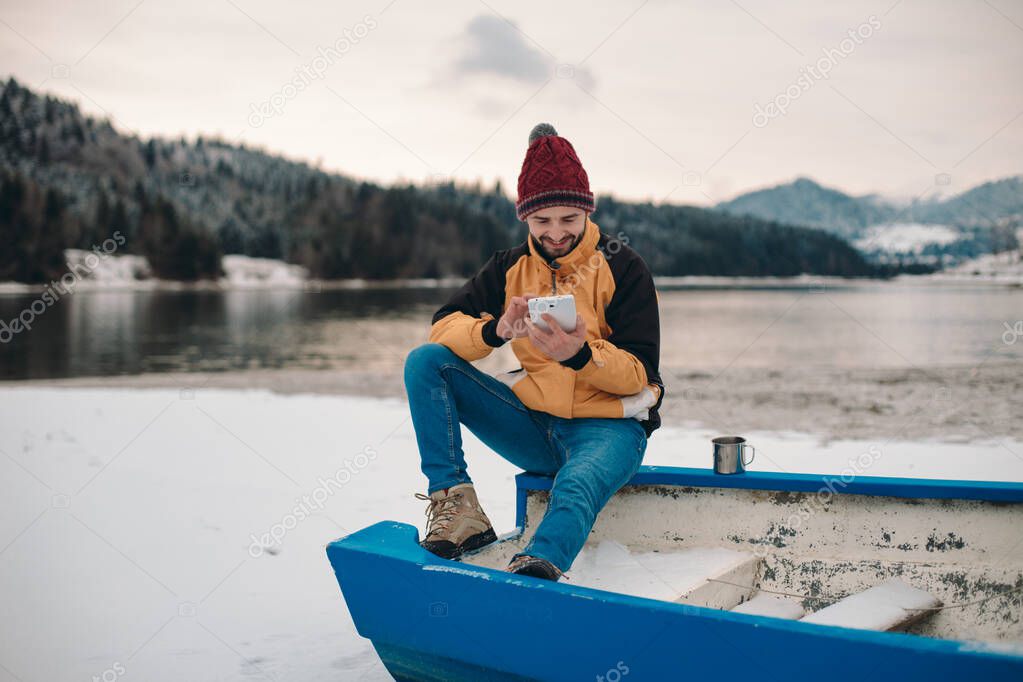 Young tourist arrived in amazing landscape place with big lake and forest , beside of a wooden house he have a little break chatting with some friends or family members using a tablet