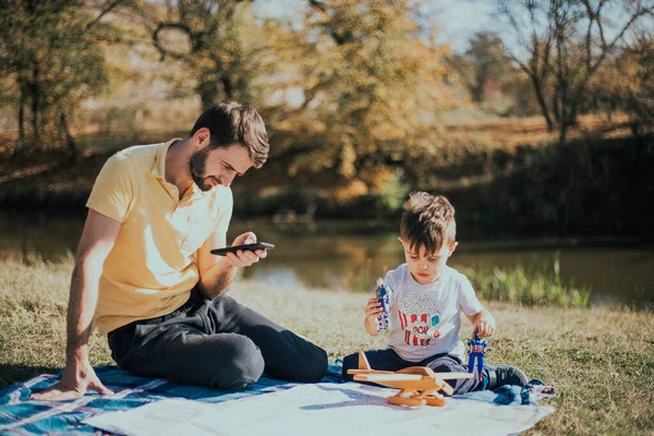 Un niño de tres años juega en un picnic cerca de su tío que mira el teléfono —  Fotos de Stock