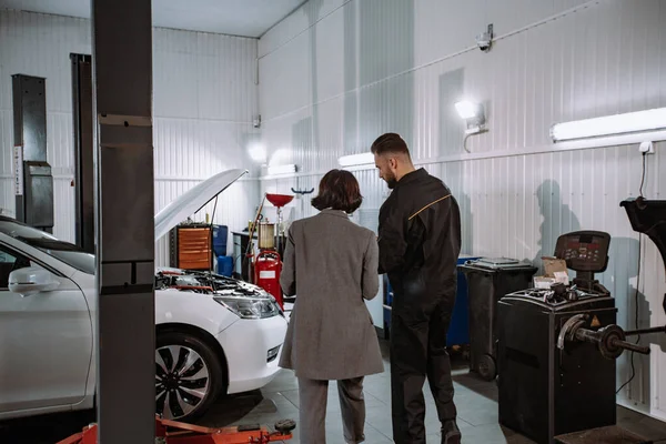 In een auto service center goed uitziende vrouw in een bedrijf pak haar naar de auto om het probleem te controleren in een auto garage — Stockfoto