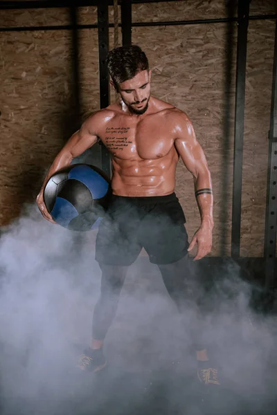Details van een atleet die een cross fit bal gebruikt om zijn zware trainingsoefeningen te doen in een donkere gymles — Stockfoto