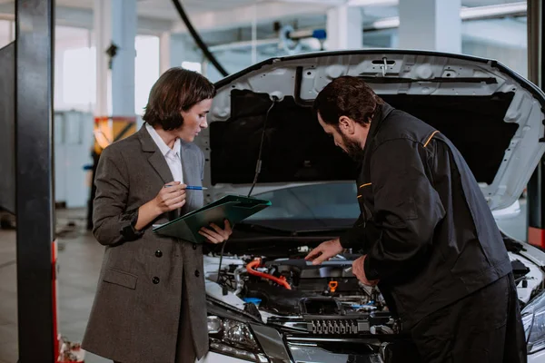 Hermosa una mujer en un traje y en servicio automático tener una conversación con el mecánico sobre el coche que muestra el problema del coche — Foto de Stock