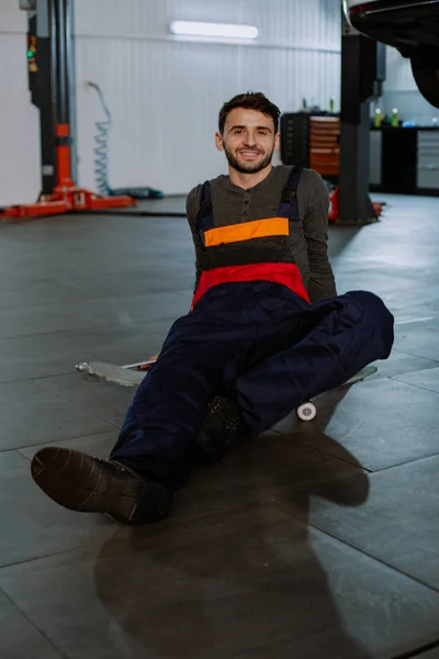 Un joven mecánico está sentado en un monopatín en un servicio de coches después de terminar el trabajo sonriendo — Foto de Stock