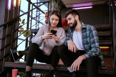 Modern çalışma stüdyosunda güzel bir erkek ve güzel bir bayan gülümsüyor merdivenlerde oturuyor ve telefondaki resimlere bakıyor.