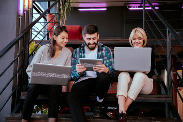 Three charismatic young office workers working hard using tablet and notebook to analyzing the plan of work while sitting on a office stairs