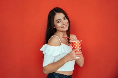 Gülümseyen genç bir kadın, elinde plastik bir bardak kola ya da gazoz tutarak stüdyoda parlak kırmızı arka planda izole edilmiş.
