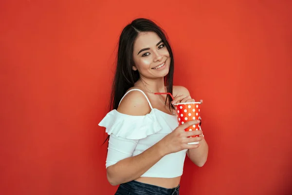 Улыбающаяся молодая женщина смотрит в сторону, держа пластиковую чашку колы или газировки изолированы на ярко-красном фоне в студии — стоковое фото
