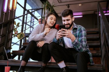 Modern çalışma stüdyosunda güzel bir erkek ve güzel bir bayan gülümsüyor merdivenlerde oturuyor ve telefondaki resimlere bakıyor.