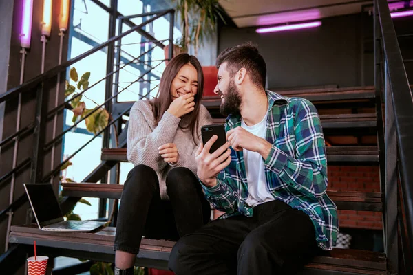 En el estudio de la oficina de trabajo moderno un chico guapo y hermosa dama sonriendo tienen un descanso sentado en las escaleras tienen, viendo un video divertido —  Fotos de Stock