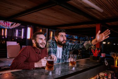 İki arkadaş bira içiyor ve barda spor müsabakaları seyrediyor.