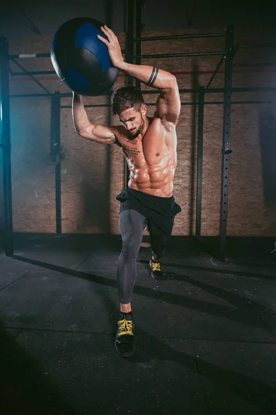 Koyu renk bir spor salonunda antrenman yapmak için çapraz formda top kullanan atletik bir adamın detayları.