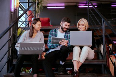 Ofis merdivenlerinde otururken not defteri ve tableti kullanarak çalışma planını analiz eden üç çekici genç ofis çalışanı.