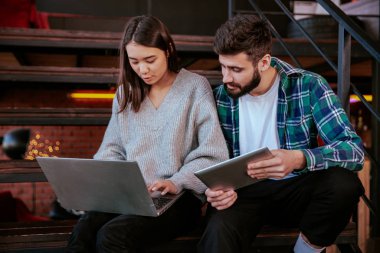 İki çekici genç ofis çalışanı ofis merdivenlerinde otururken not defteri ve tableti kullanarak çalışma planını analiz ediyorlar.
