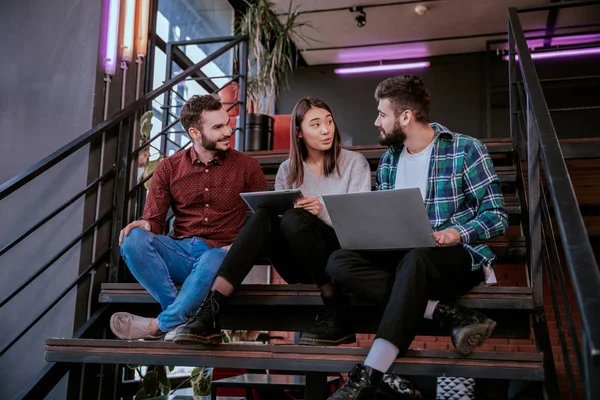 In a modern office stairs three attractive workers two guys and one lady analyzing the plan of work while sitting on the stairs