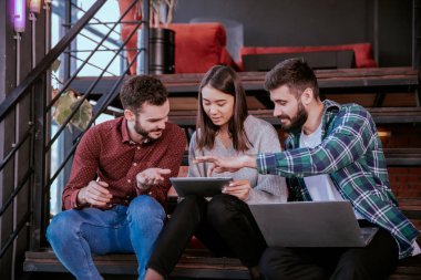 Üç karizmatik genç ofis çalışanı ofis merdivenlerinde otururken çalışma planını analiz etmek için tablet ve not defteri kullanarak çok çalışıyorlar.