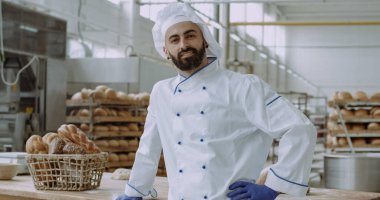 Bir fırın mutfağında çalışma masasının yanında dururken şık bir üniformayla gülümseyen yakışıklı bir yüz fırıncısının kamera önünde fotoğrafı.