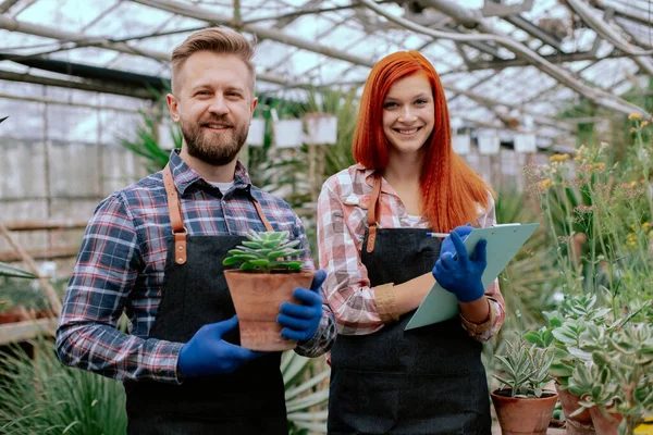 Beautiful redhead florist and an attractive gardener in his own flower greenhouse smiling large looking straight to the camera smiling large