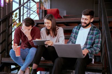Üç karizmatik genç ofis çalışanı ofis merdivenlerinde otururken çalışma planını analiz etmek için tablet ve not defteri kullanarak çok çalışıyorlar.