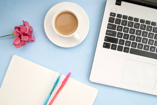 Concept workplace. Laptop keyboard, white cup of coffee with milk, open notebook for notes, ballpoint pen, pink flower on a blue background, place for text, copy space. Top view. Flat lay