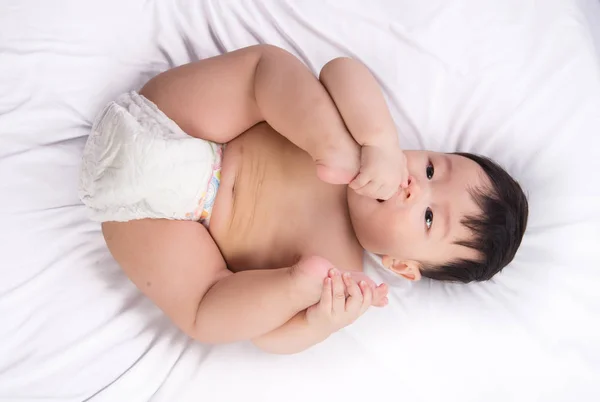 Retrato de lindo niño asiático de 6 meses de edad poner los pies en la boca en la cama blanca —  Fotos de Stock