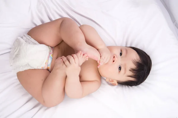 Retrato de lindo niño asiático de 6 meses de edad poner los pies en la boca en la cama blanca — Foto de Stock