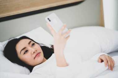 Asyalı kadın yatakta cep telefonuyla yazıp sohbet ediyor.