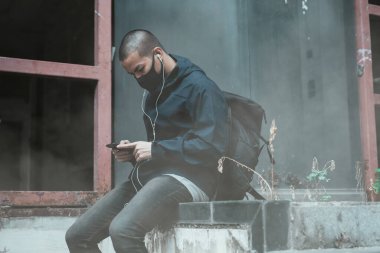 Şık bir adamın portresi, tasasız bir şekilde dışarıda maskeyle oturuyor, şehirde uzun bir yürüyüşten sonra dinleniyor, modern cep telefonunu eğlence için kullanıyor, kulaklıkla rock müzik dinliyor.