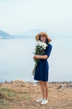 Uzun boylu Taylandlı güzel bir kız gölün kenarında beyaz çiçekleri kucağında kameraya bakıyor..
